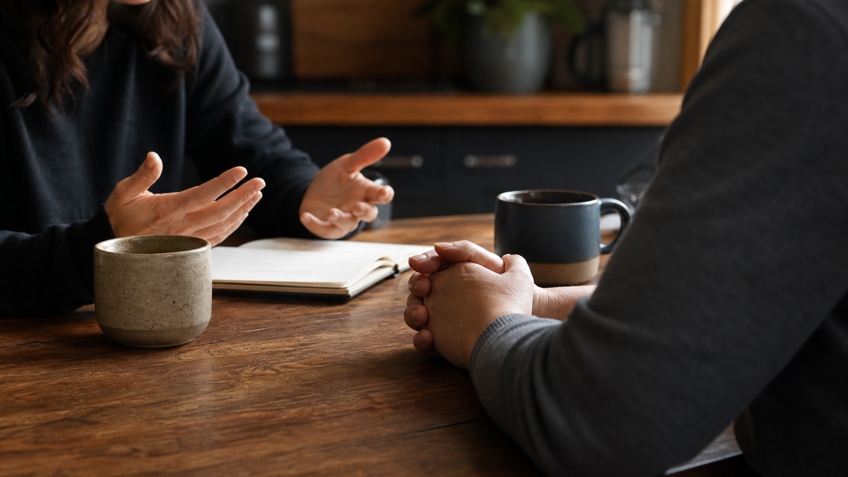 Two adults having a calm conversation at a kitchen table with a notebook and mugs